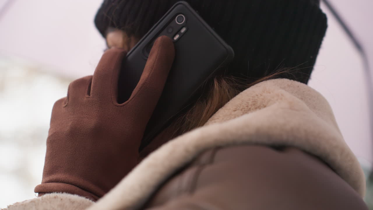 Close-up side view of lady in black cap, brown jacket, and gloves, holding black phone to her ear, with a blurred background of nature and urban elements