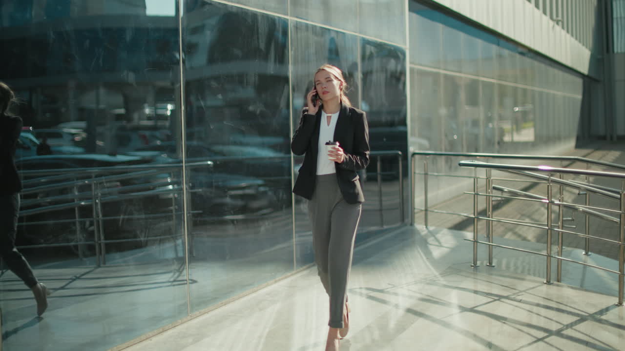 Young elegant banker woman walking confidently with coffee in hand while on phone call near modern glass building, exuding professionalism and focus as she strolls through bright urban environment