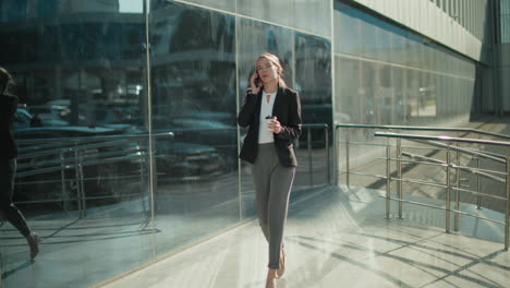 young elegant banker woman walking confidently with coffee in hand while on phone call near modern glass building, exuding professionalism and focus as she strolls through bright urban environment