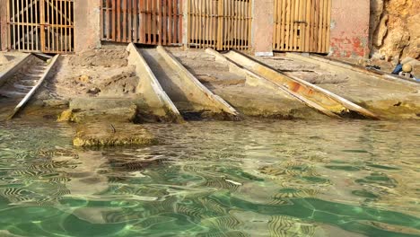 Viejos-Cobertizos-De-Barcos-De-Pesca-Junto-Al-Mar-En-Ibiza-España,-Cabañas-De-Pesca-Tradicionales-En-Una-Playa-Soleada,-Garajes-De-Barcos-En-La-Orilla-Con-Agua-Turquesa,-Tiro-De-4k