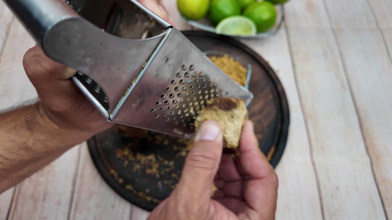 close-up authentic manual process of hands grating a block of hard panela (raw sugar cane) with a metal grater over a wooden board. The scene highlights fresh limes in the background