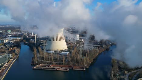 aerial view of a thermal power plant emitting smoke on a sunny day, with a river and a city in the background. beautiful aerial view flight ascending drone