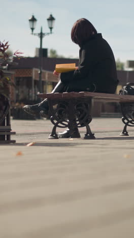 back view of white girl seated in public space, focused on her book, with flower pot swaying gently in wind and pedestrians walking in background, stores with inscription visible across the road