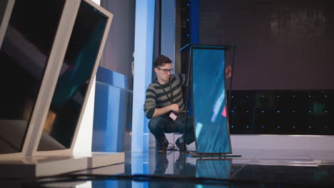 man adjusts digital display screen setup in video production studio, focused on configuring podium, with camera setup in background, blue lighting, preparing studio space for filming and broadcasting