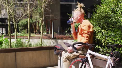 hombre afroamericano albino pensativo con rastas sentado en el parque bebiendo café, usando una computadora portátil