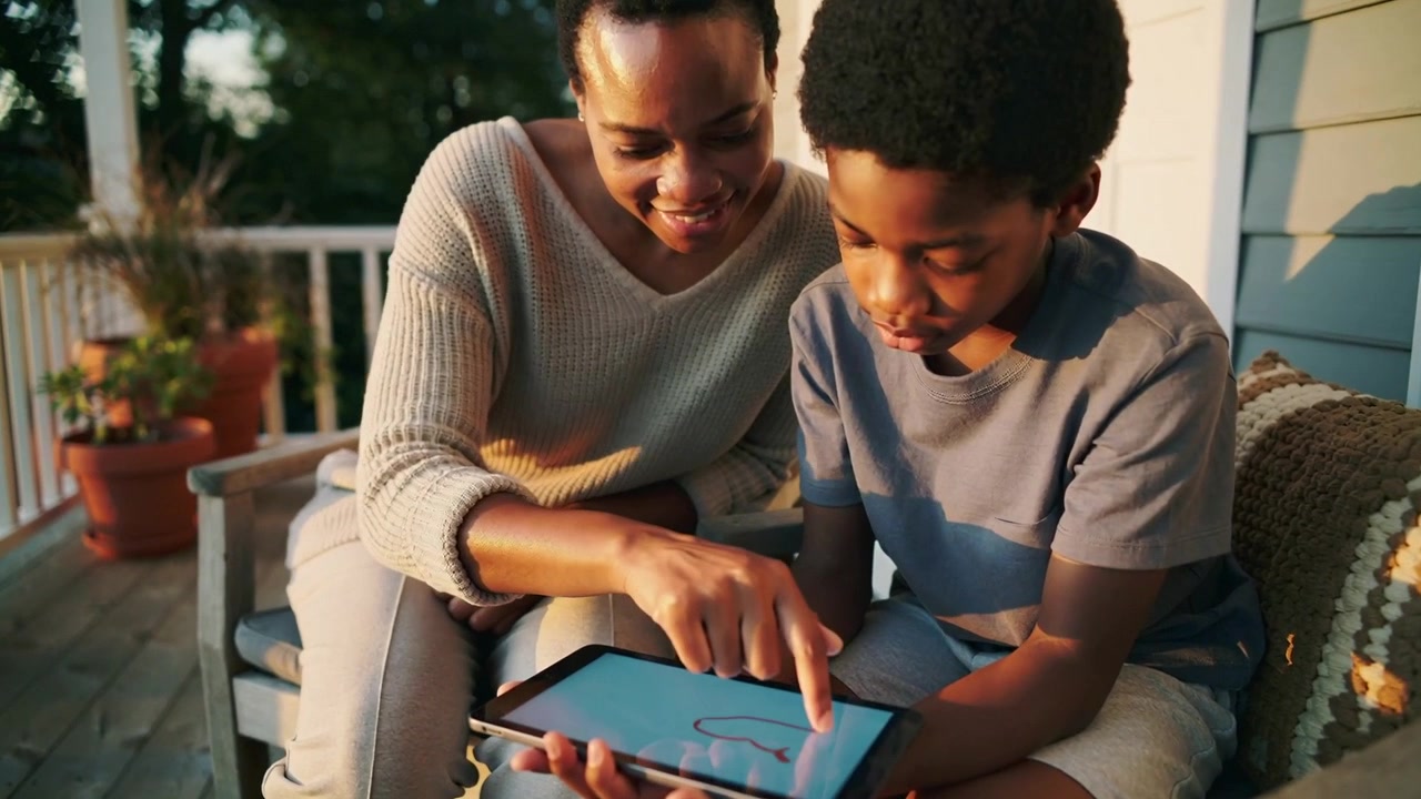 Mère pointant du doigt, se penchant pour apprendre à son fils à dessiner sur un banc de porche, traçant un cœur sur l'écran tactile d'une tablette.