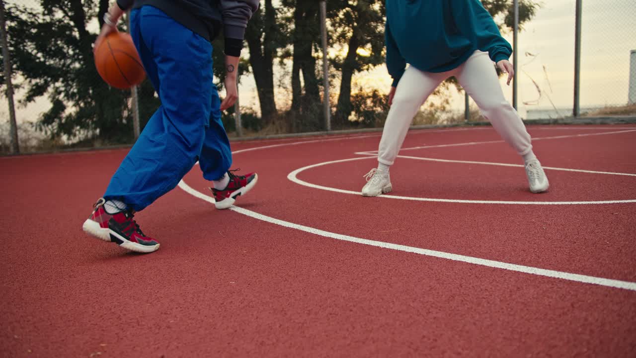 Close-up shot of a girl in blue pants maneuvering and hitting the ball off the floor while playing basketball with a girl in white pants on a red basketball court early in the morning