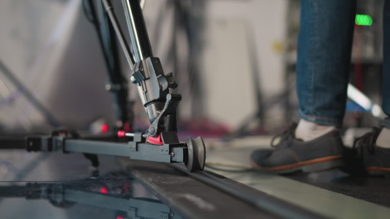 Close-up shot of camera dolly system on track in professional studio. Camera rig is in motion as technician adjusts gear. Blurred background with studio lighting and colorful equipment visible