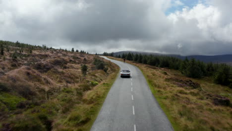 Forwards-tracking-of-sports-car-passing-against-cyclist-riding-downhill.-Road-in-highlands-landscape-under-cloudy-sky.-Ireland