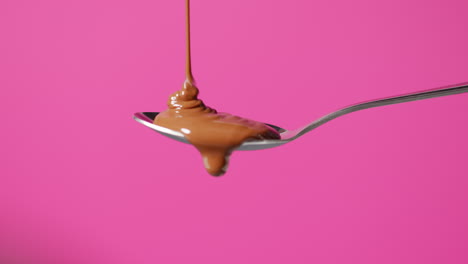 close up of melted chocolate being poured onto spoon and overflowing against pink coloured background with copy space