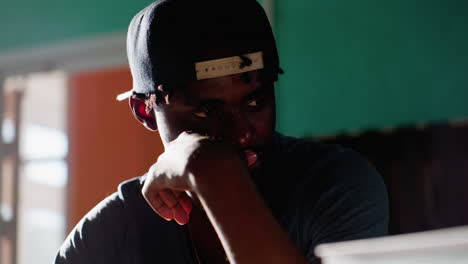 close up reflective man wearing cap in library studying at table with books and notes, resting chin on hand while lost in thought, soft light on face, teal wall and shelves behind