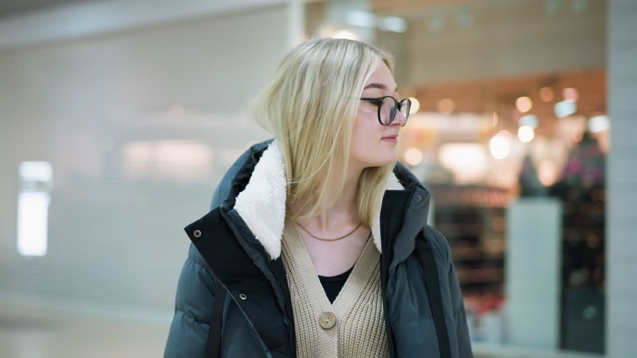 Beautiful lady in glasses walking through well-lit mall, looking contemplative, blurred bokeh light effect seen through glass with reflections, soft focus on background featuring indistinct objects