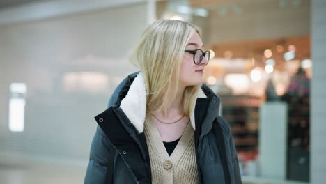 beautiful lady in glasses walking through well-lit mall, looking contemplative, blurred bokeh light effect seen through glass with reflections, soft focus on background featuring indistinct objects