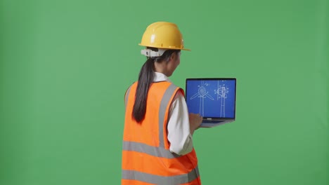 back view of asian female engineer with safety helmet looking at the wind turbine blueprint on laptop while standing in the green screen background studio