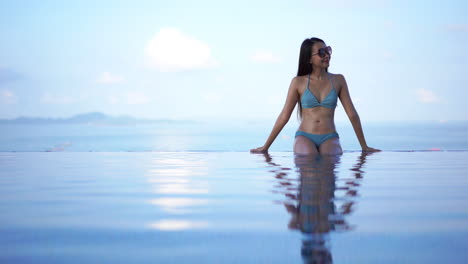 Bonita,-En-Forma,-Joven-Mujer-Asiática-En-Bikini-Azul-Sentada-En-El-Borde-De-Una-Piscina-Infinita-Disfrutando-Del-Cálido-Sol-Tropical