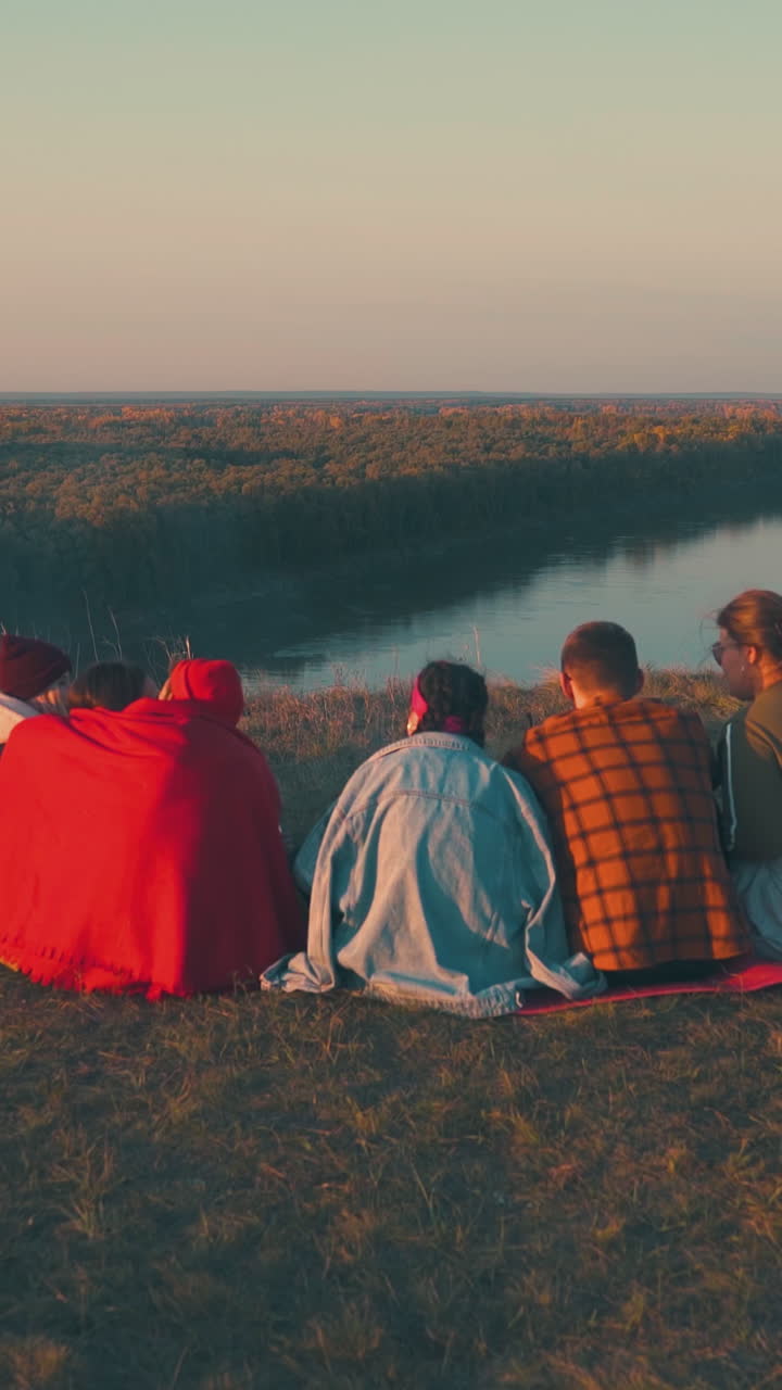 happy tourist group rests at burning bonfire on steep river bank against fantastic autumn landscape at sunset backside view