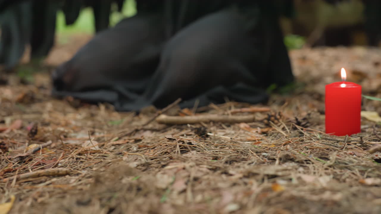 Close up of ritual scene in forest with four glowing red candles arranged on ground while person kneels nearby, creating atmosphere of mystery, devotion, and sacred spiritual energy