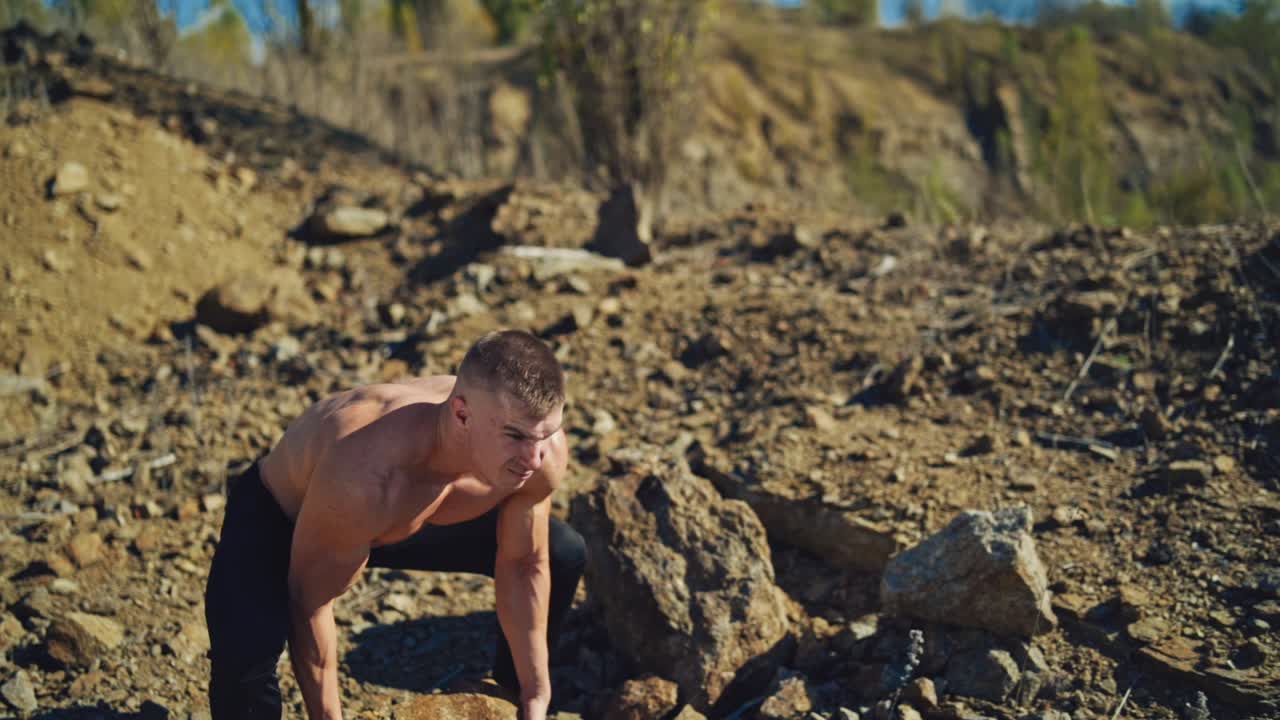 Athletic man exercises with stones. Strong sportsman taking heavy stone and throwing it over the head behind the back on rocky background. Slow motion.