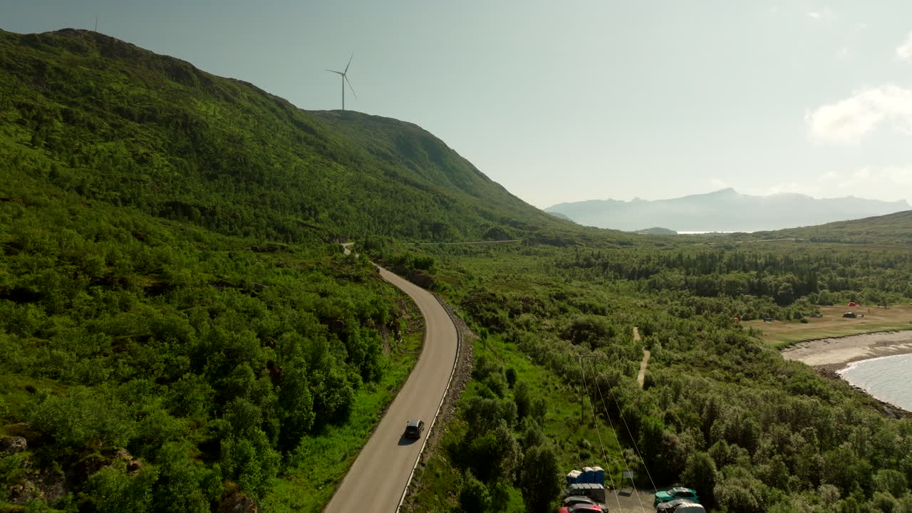 Car on winding road through lush green landscape, Wind turbine on hill, Kvaløya, Troms, Norway. Aerial forward