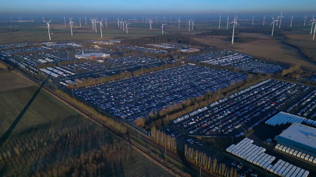 used car trade awaiting transport at a large distribution center with warehouses and railway tracks, surrounded by wind turbines. Unique aerial view