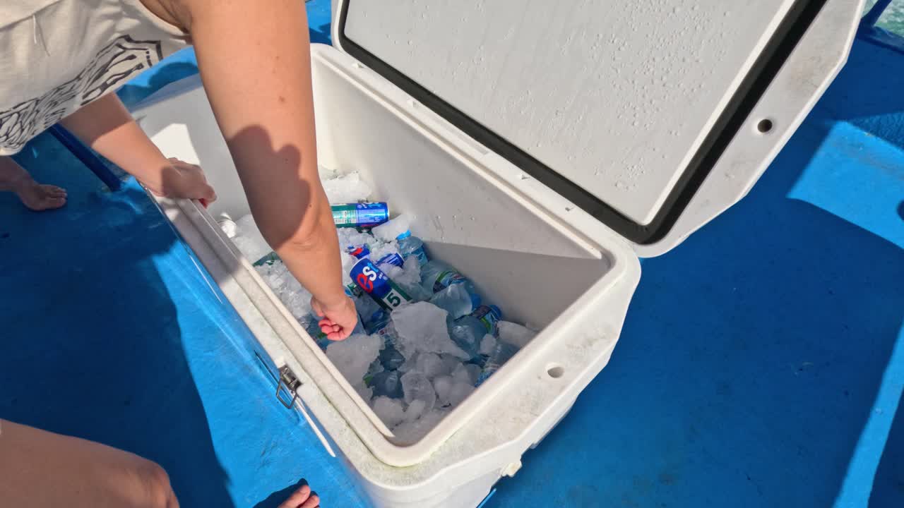 A person arranges bottled drinks in an ice-filled cooler under bright sunlight on a boat in Phuket, Thailand