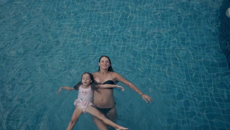young-woman-holds-floating-little-girl-in-blue-pool-top-view