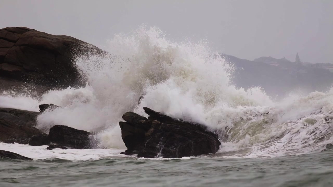 Violent ocean swell colliding with a large rock formation, generating massive white water spray and foam. Fixed tripod telephoto shot. Overcast sky with hazy coastal hills in the background.