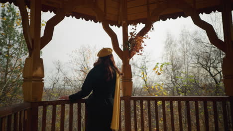 elegant lady in yellow beret and muffler explores a wooden gazebo, pausing to place her hand on the railing, looking out pensively amid the autumn foliage backdrop