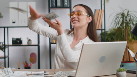 cheerful rich business woman working on office laptop wasting throwing money to camera profit win