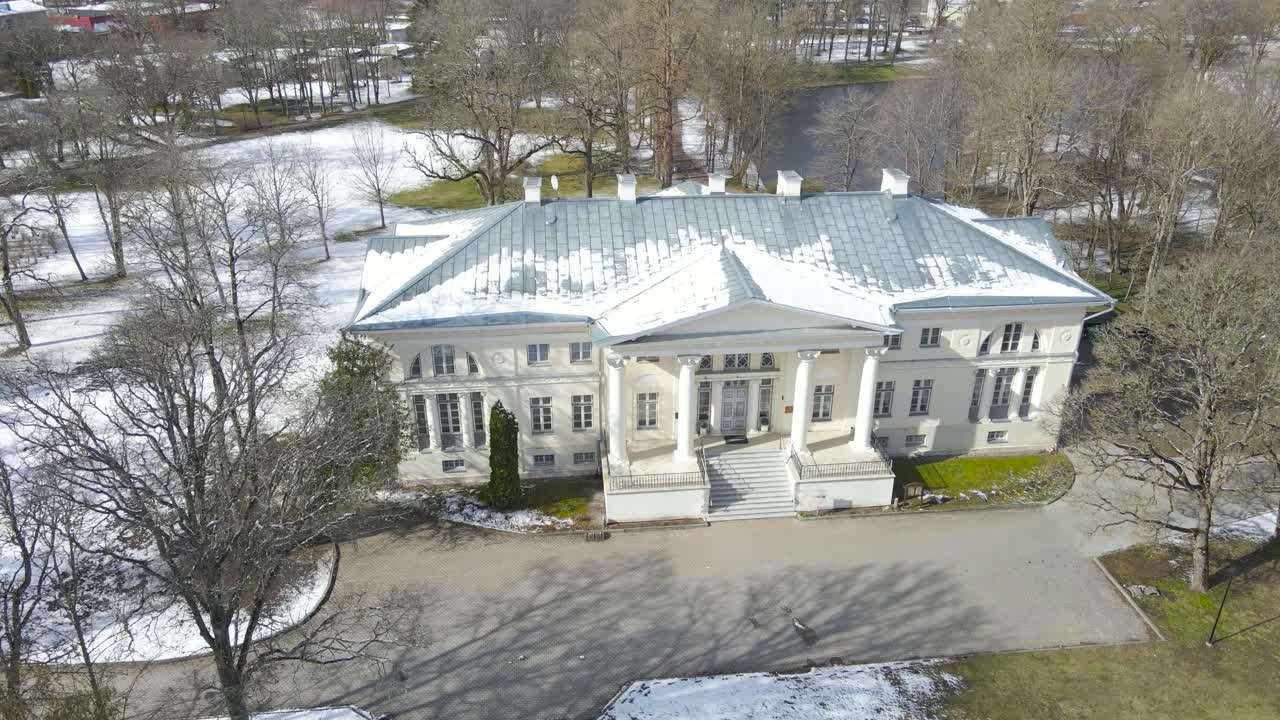 Aerial drone footage orbiting and spinning around a gorgeous large white mansion or manor called Saku mõis during winter sunny day while green mowed lawn grass visible in the garden and leafless trees