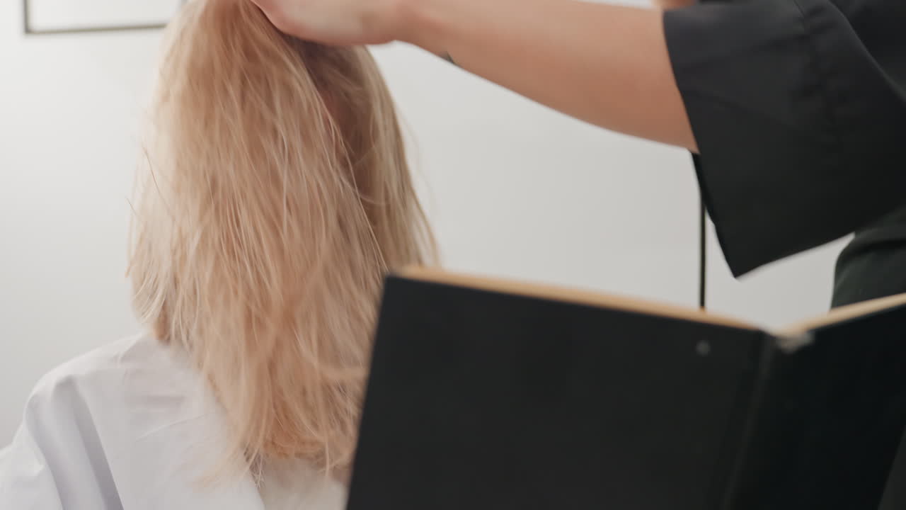 Primer plano desde atrás de una mujer caucásica con pelo rubio encrespado cubriéndole la cara sentada con una capa de peluquería mientras el estilista levanta el secador que muestra mechones enredados y textura indomable, escena de preparación espontánea, suave natural
