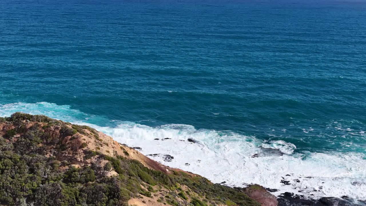 Aerial camera pans above Cape Schanck cliffs, revealing blue ocean waves and rugged shoreline