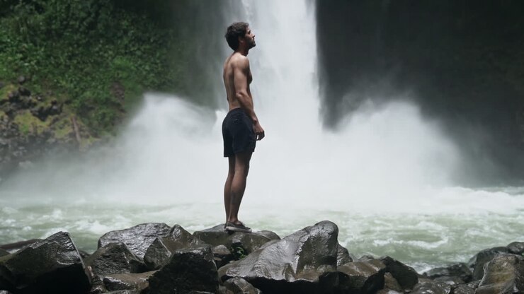 Man looks around as jungle waterfall crashes down behind him, slow motion