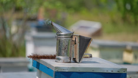A metal smoker with white smoke coming from it and a brush at the beehive. Instrument and tool for apiculture work. Blurred backdrop.