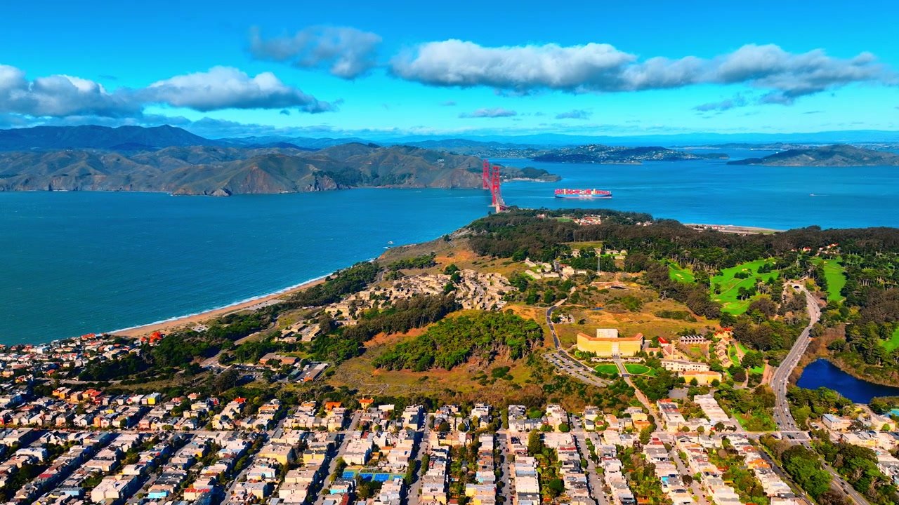 Flyfoto av Golden Gate Bridge og Presidio parkområde, San Francisco. Droneperspektiv ovenfra av kystklipper, lasteskip og grønne skogkledde åser.