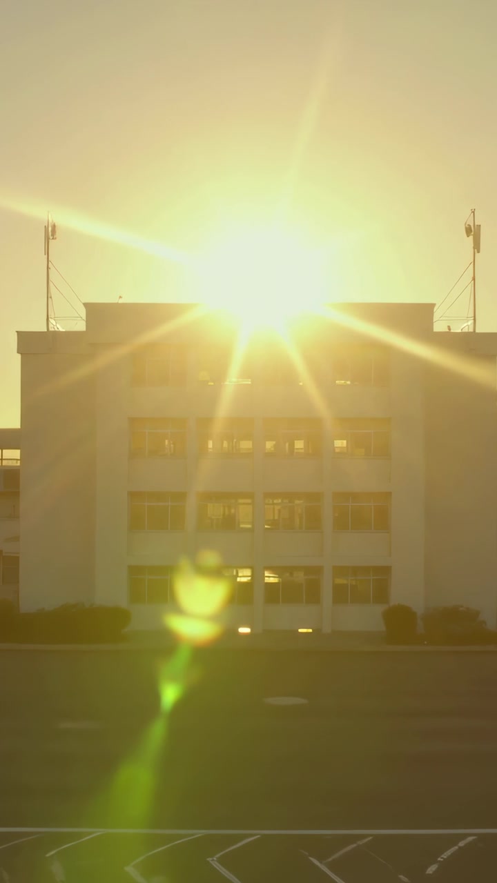 Vídeo vertical: Câmera enquadrando um edifício de escritórios enquanto o sol acima do telhado projeta um reflexo de lente verde no estacionamento.