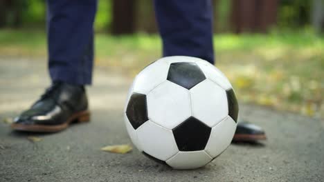 close-up male foot in elegant leather shoe step on soccer ball outdoors. unrecognizable stylish businessman with football equipment in park. lifestyle and hobby.