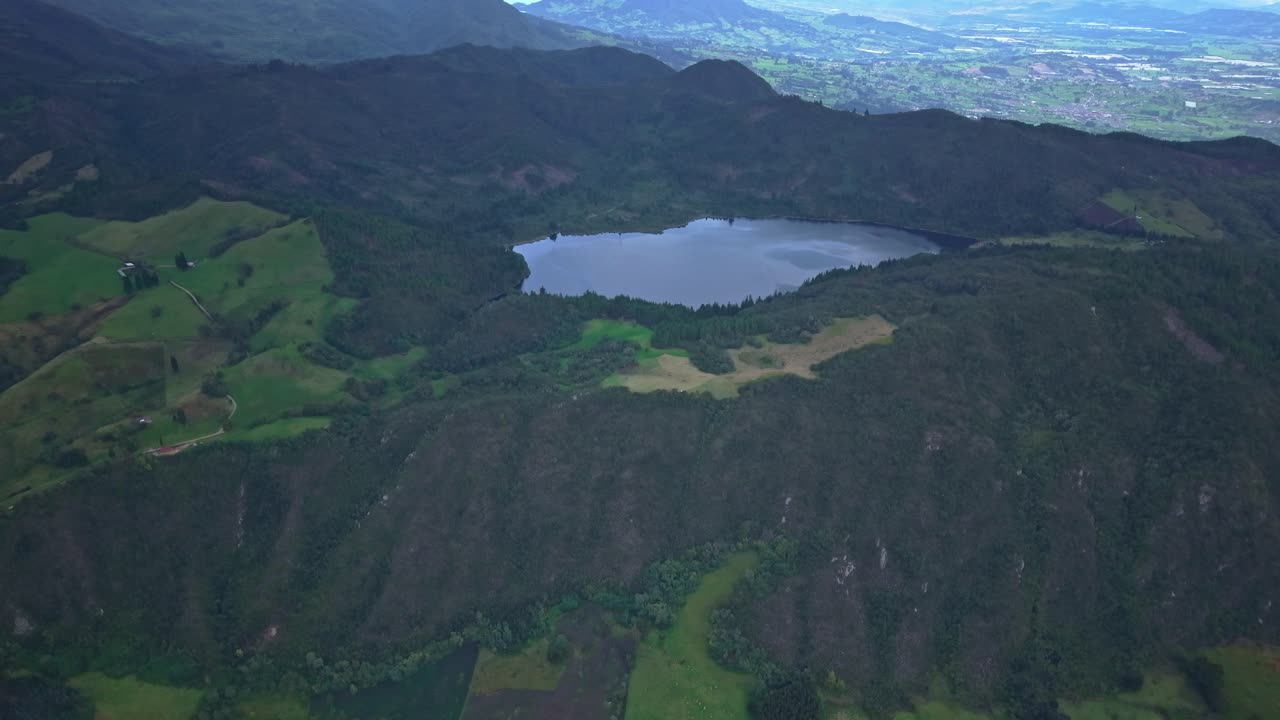 Drone rises above Laguna de Pantano Redondo in Zipaquirá, revealing the surrounding volcanic slopes and green farmland beneath soft daylight