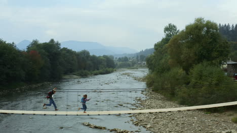 Junges-Paar-Läuft-über-Eine-Holzhängebrücke-über-Einen-Fluss-Im-Wald