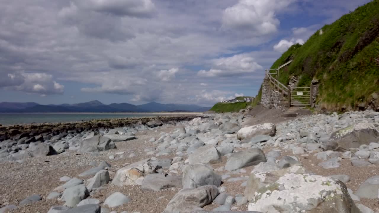 영국의 웨일즈에서 먼 산이 있는 어두운 바위까지 바위가 많은 해변을 가로질러 오른쪽에서 왼쪽으로 느린 드론 트랙 - 10초 버전 2