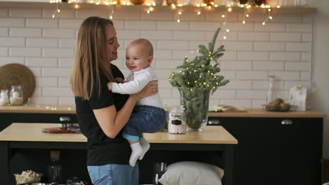 mom hugs the baby and the child smiles looking at his beloved mother. together stand in the white kitchen on christmas eve on the background of garlands and christmas trees. happy mother and baby kissing and hugging.maternity concept motherhood beautiful happy family stock video footage in slow moti