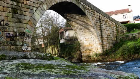 Vista-En-ángulo-De-La-Luz-Del-Sol-Sobre-Un-Puente-De-Ladrillo-Con-Rocas-Cubiertas-De-Líquenes-Cubiertos-De-Musgo-Junto-Al-Río-Lonia-En-Ourense