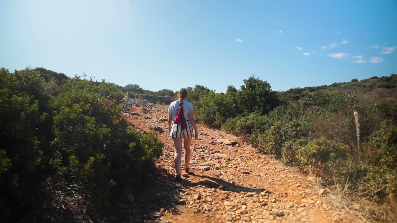 White brunette with fuschia scarf braided in her hair walkin with shorts on a dirt path surrounded by low bushes.