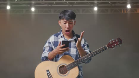 asian teenager with guitar using smartphone and saying wow while standing on stage