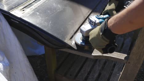 angle grinder being used to grind rust away on old black car door that's resting on wooden chairs in summer daytime