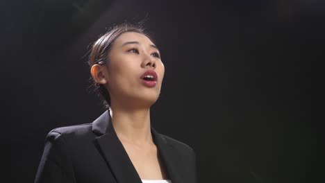 side view of asian speaker woman in business suit showing index fingers up while speaking in the black screen studio