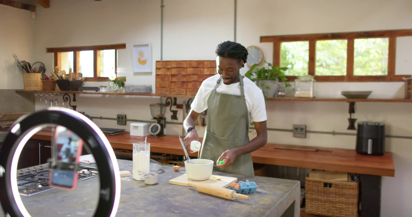 Uomo afroamericano adulto che sbatte con una frusta all'isola della cucina, con una ring light che filma una ricetta, versando il latte.