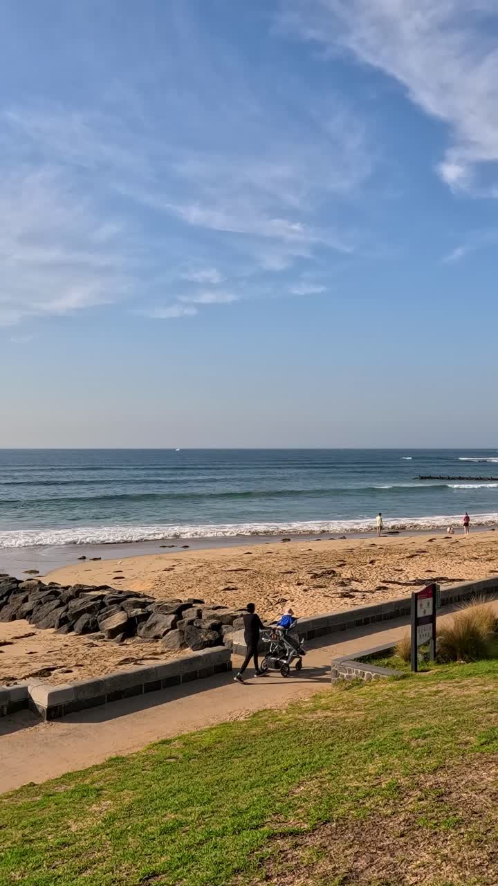 Two cyclists ride along a path by the beach under clear skies, capturing a serene coastal moment