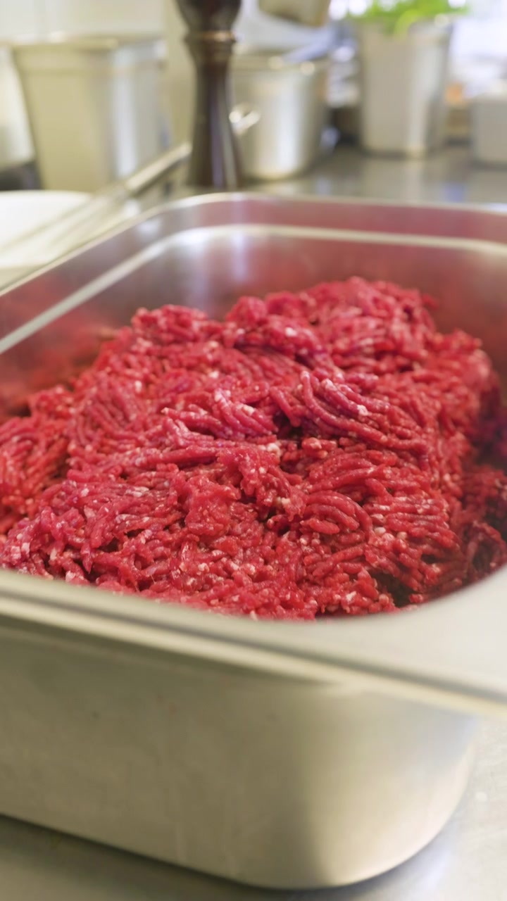 Handheld pull out shot of fresh raw minced meat in a stainless steel kitchen tray, filmed in a professional food preparation environment.