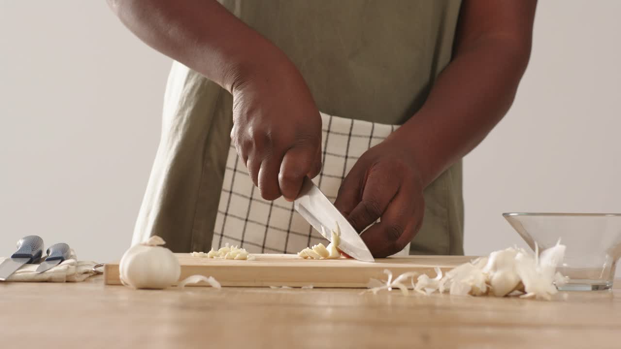 Uomo afroamericano che tiene uno spicchio d'aglio, lo sbuccia e lo trita su un tagliere con un coltello, preparando un pasto.